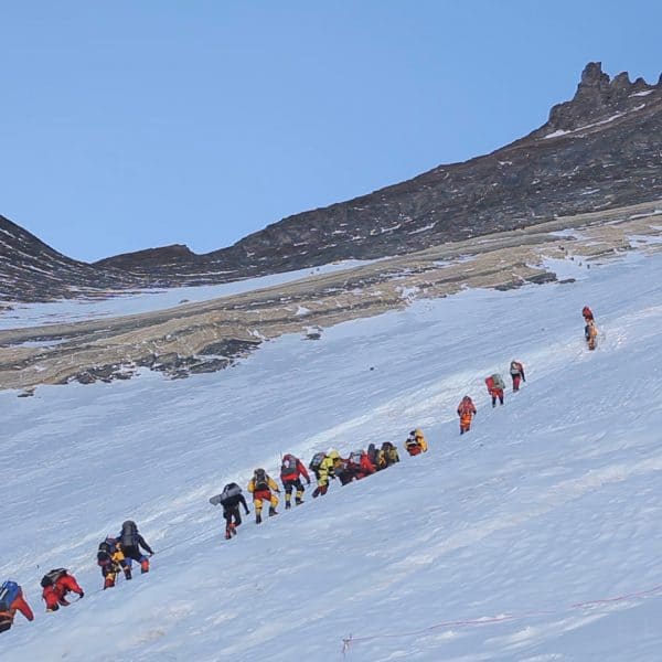Line up of climbers in multi colored suits climbing an icy section on a mountain with yellow rock protruding from the ice with jagged rocks high above