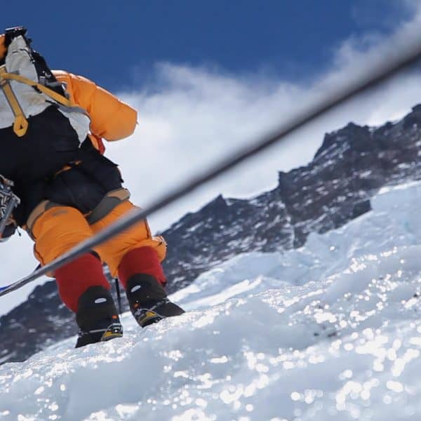 A climber in an orange down suit seen from behind using a sagety line to climb a steep icy face with mountains in the distance
