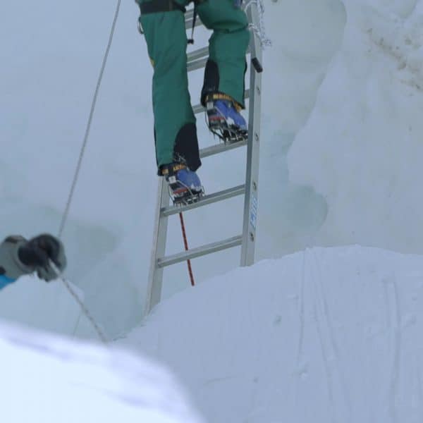 A man holds a rop as legs can be seen climbing down an aluminum ladder placed in the middle of a glacier next to a wall of ice