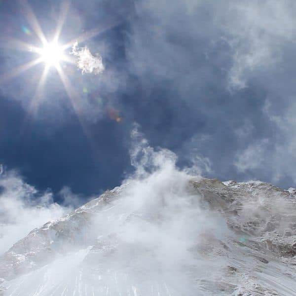 A snow capped summit from a low angle surrounded by clouds with the bright sun shining from above