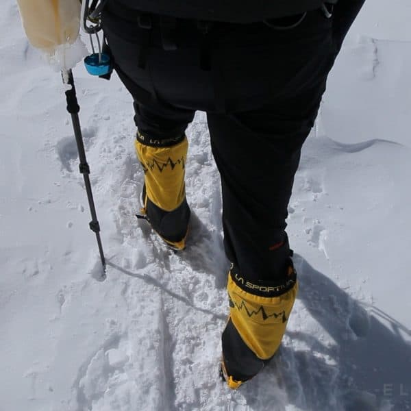 A climbers legs and yellow boots are seen while walking on a snowy glacier with a ski pole
