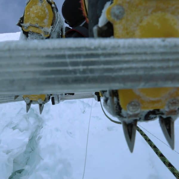 A climber climbs an aluminum ladder with crampons with snow and a visible mountain int he distance