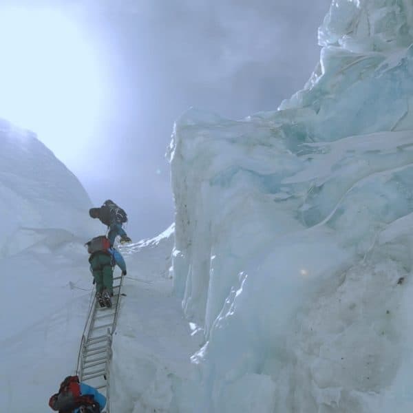 Three men climb down a dangerous ladderssecured against a giant wall of ice with the bright sun shining from above