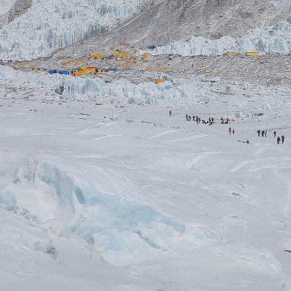 Climbers are seen walking on a snoy path with dozens of yellow tents placed on a rocky and snowy glacier in the distance