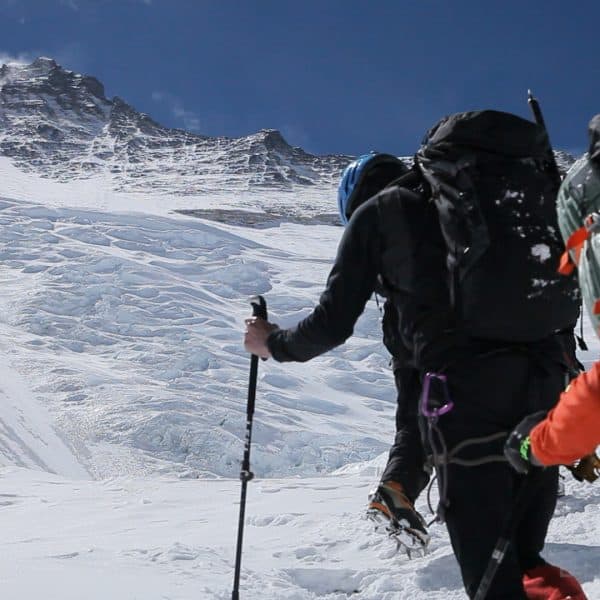 Two climbers walk with ski poles on a glacier towards a steep icy face of a tall mountain called Lhotse