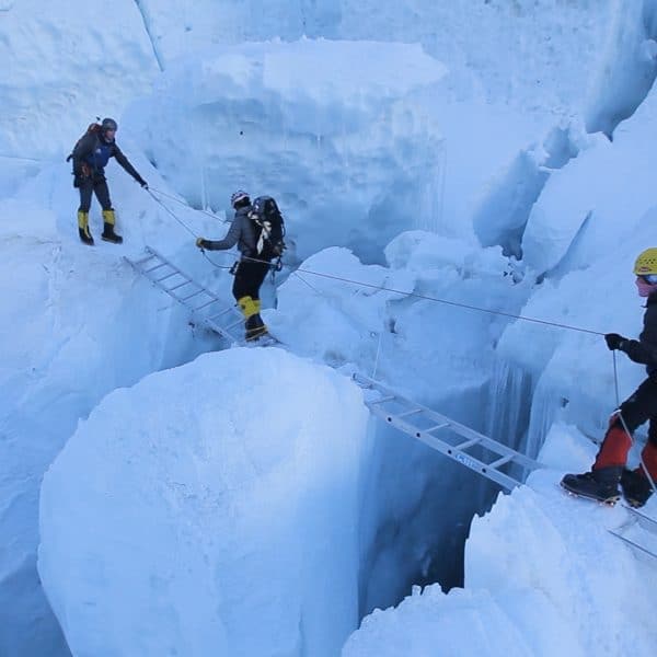 2 climbers assist a man in grey jacked crossing an aluminum ladder suspended over a crevasse on a dangerous ice field