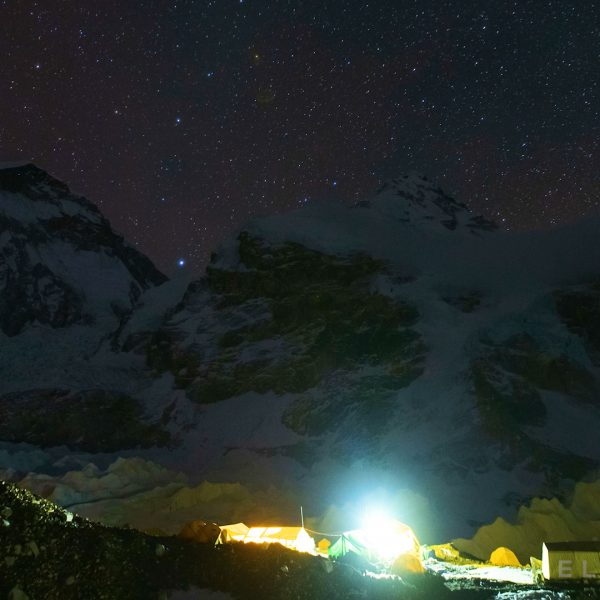 Two mountains tower above multiple yellow tents at night with a purpulish starry sky