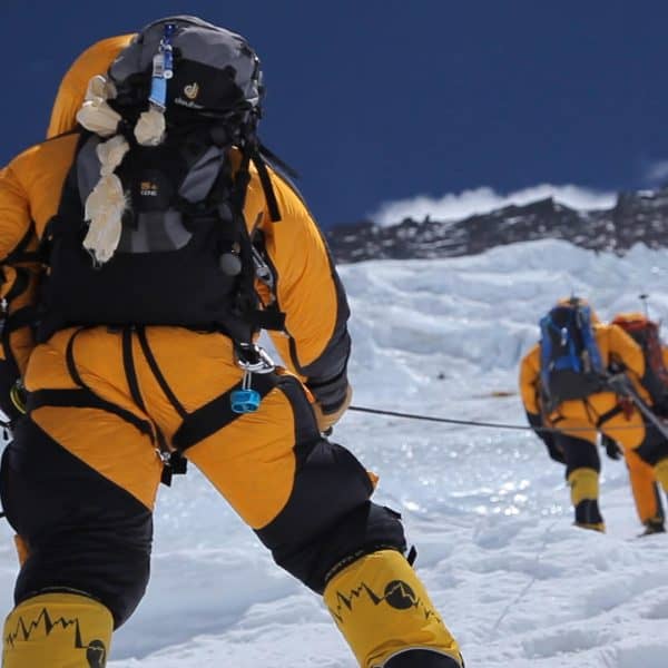 Three climbers walk in crampons dressed in warm yellow suits up a steep icy face with a tall mountain called Lhotse in the distance