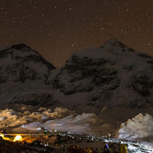 Tents are set up on a snowy rocky glacier at night with two mountains in the background and a sky filled with stars