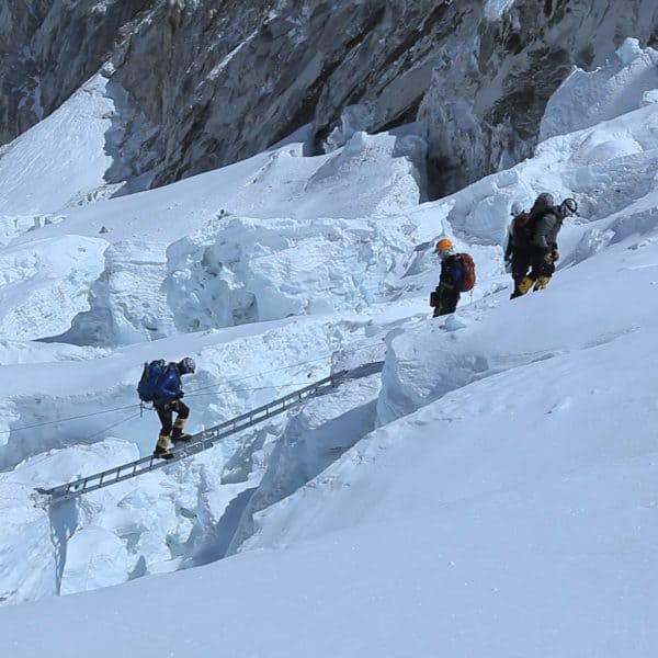 5 climbers in a dangerous snowy landscape as one dressed in blue crosses an aluminum ladder suspended over a crevasse