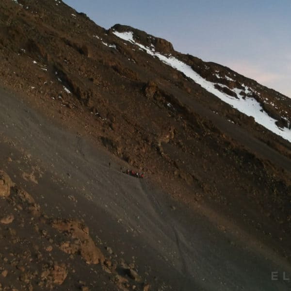 A team of climbers walk a steep narrow path at sunrise with snow in the distance