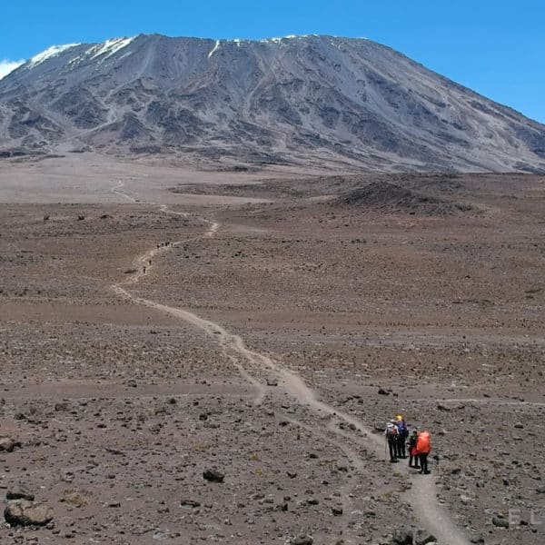 A large volcano like mountain towers above a dry path and open valley with a crystal clear blue sky as climbers follow a path to the summit