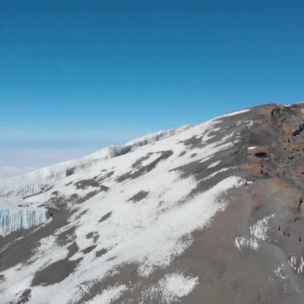 Climbers walk down the summit of Kilimanjaro mountain on a dry rocky trail with some visible snow and a glacier in the background and large boulder in the foreground