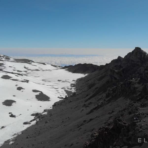 A snowy lanscape contrasted with dark dry rocky terrain with a path used by trekkers on Kilimanjaro