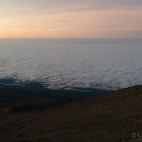 A sea of clouds with an orangish sky with dark brown terrain on the side of a mountain